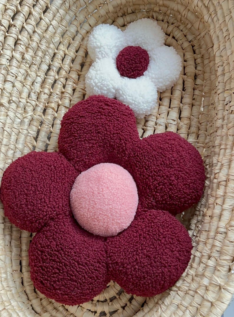 May include: Two flower-shaped decorative pillows. One is white with a burgundy center, and the other is burgundy with a pink center. The pillows have a soft, textured appearance and are placed inside a woven basket.