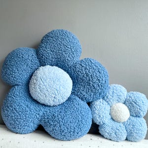 May include: Two plush flower-shaped pillows in shades of blue. The larger pillow has a light blue center, while the smaller pillow has a white center. The pillows are made of a soft, textured material, perfect for a nursery or bedroom.