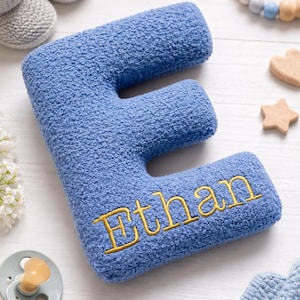 May include: A plush, blue, textured letter E cushion featuring the name Ethan embroidered in gold thread across the front. The soft, fuzzy fabric has a distinct looped pile surface, and the letter is shaped in a bold, block-style font.