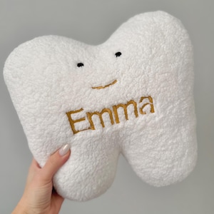 Può includere: Un cuscino in peluche bianco con texture tipo sherpa a forma di dente. Il cuscino presenta due piccoli occhi ricamati in nero e un semplice sorriso ricamato in oro. Il nome Emma è ricamato con filo dorato al centro del dente.