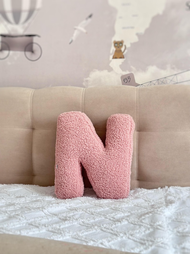 K&ouml;nnte beinhalten: Ein buchstabf&ouml;rmiges Dekokissen in Form des Buchstabens N. Das Kissen ist mit einem strukturierten, altrosa Sherpa- oder Boucl&eacute;-Stoff bezogen, der ihm ein weiches, flauschiges Aussehen verleiht. Es ist ein eigenst&auml;ndiges Dekorationsobjekt mit abgerundeter Form.