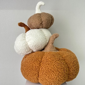 Könnte beinhalten: Ein Stapel aus drei Plüschkürbissen in Herbstfarben. Die Kürbisse sind in Orange-, Weiß- und Brauntönen gehalten und haben strukturierte Oberflächen und gebogene Stiele. Ein herbstliches Dekorationsstück.
