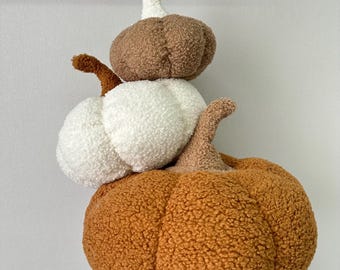 May include: A stack of three plush pumpkins in shades of orange, white, and brown. The pumpkins have textured, fuzzy surfaces and curved stems. The arrangement is a decorative fall-themed item.