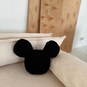 May include: A black, plush Mickey Mouse-shaped pillow rests on a beige sofa. The pillow has a rounded head and two large, circular ears. The background includes a wooden panel and a white wall.