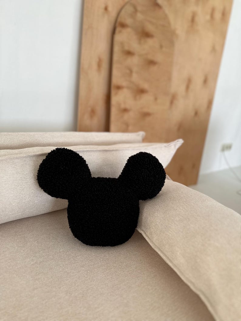 May include: A black, plush Mickey Mouse-shaped pillow rests on a beige sofa. The pillow has a rounded head and two large, circular ears. The background includes a wooden panel and a white wall.