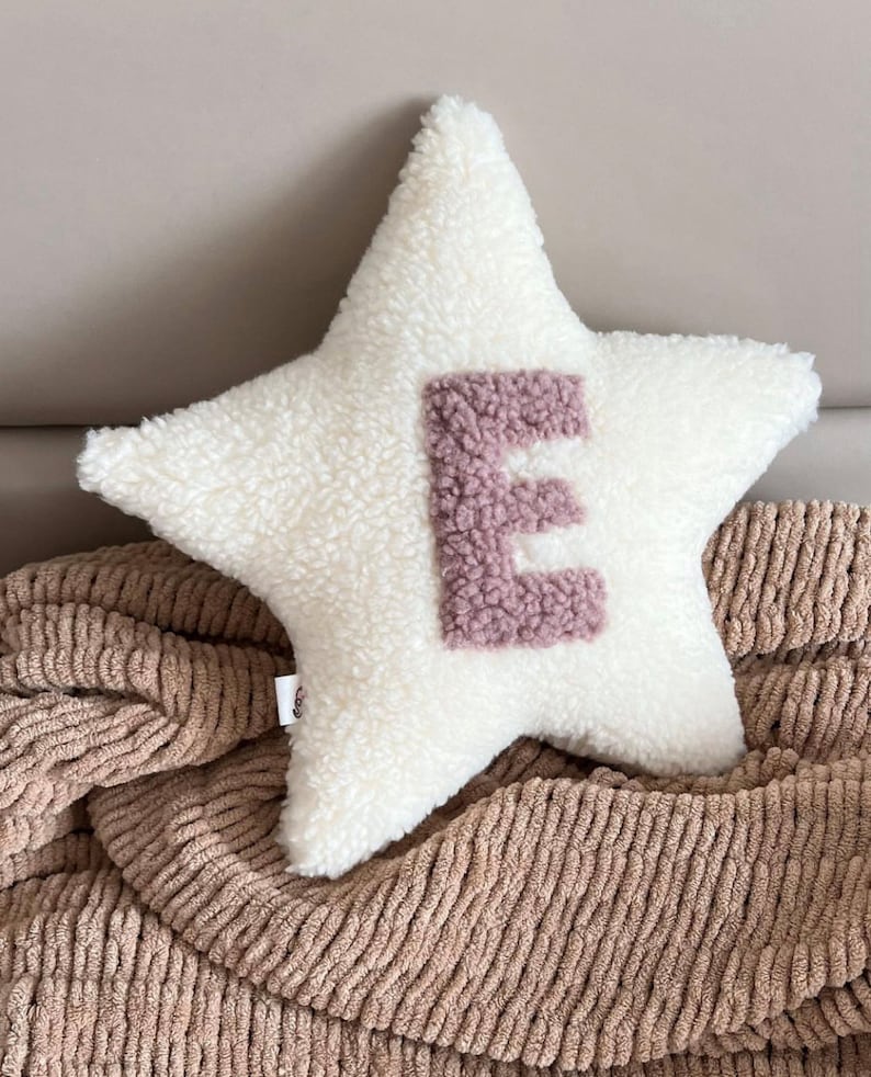 以下が含まれることがあります： 中央にピンクのふわふわの文字「E」がついた白い星型のクッション。クッションは茶色のニットの毛布の上に置かれています。