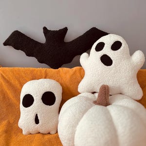 May include: A set of four plush Halloween-themed pillows. The pillows are shaped like a bat, a ghost, a pumpkin, and a skull. The pillows are all white with black accents.