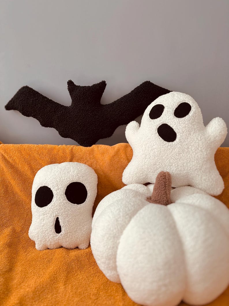 May include: A set of four plush Halloween-themed pillows. The pillows are shaped like a bat, a ghost, a pumpkin, and a skull. The pillows are all white with black accents.
