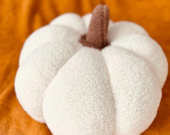 May include: A white plush pumpkin with a brown stem. The pumpkin is sitting on a fuzzy orange surface.