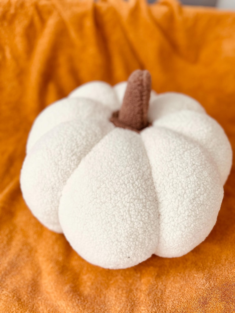 May include: A white plush pumpkin with a brown stem. The pumpkin is sitting on a fuzzy orange surface.