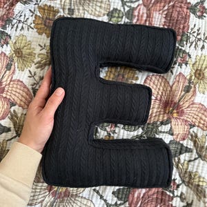 May include: Black knitted letter E cushion. The cushion is made of a soft, textured fabric and is shaped like the letter E.