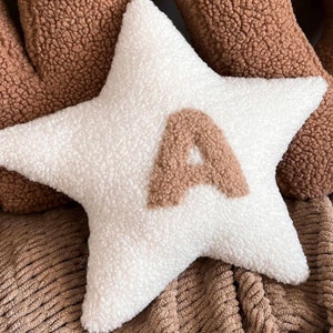 以下が含まれることがあります： 中央に茶色のふわふわした文字「A」がついた白い星型のクッション。クッションは茶色の質感のある表面の上に置かれています。