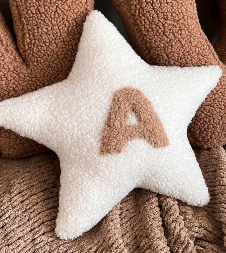 以下が含まれることがあります： 中央に茶色のふわふわした文字「A」がついた白い星型のクッション。クッションは茶色の質感のある表面の上に置かれています。
