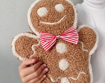 May include: A gingerbread man cushion with a brown, textured body, white frosting trim, and a red and white striped bow tie. The cushion features a smiling face with white button details. A festive Christmas decoration.