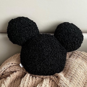 Puede incluir: Un cojín negro texturizado con forma de cabeza y orejas de Mickey Mouse. El cojín está hecho de un material suave y afelpado y descansa sobre una manta beige tejida. Las orejas son redondas y la cabeza es ovalada.