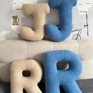 K&ouml;nnte beinhalten: Vier Buchstabenkissen aus strukturiertem, sherpa-artigem Stoff. Zwei Kissen haben die Form des Buchstabens J, eines in Beige und eines in Blau. Zwei Kissen haben die Form des Buchstabens R, eines in Beige und eines in Blau. Weiche, abgerundete Form.