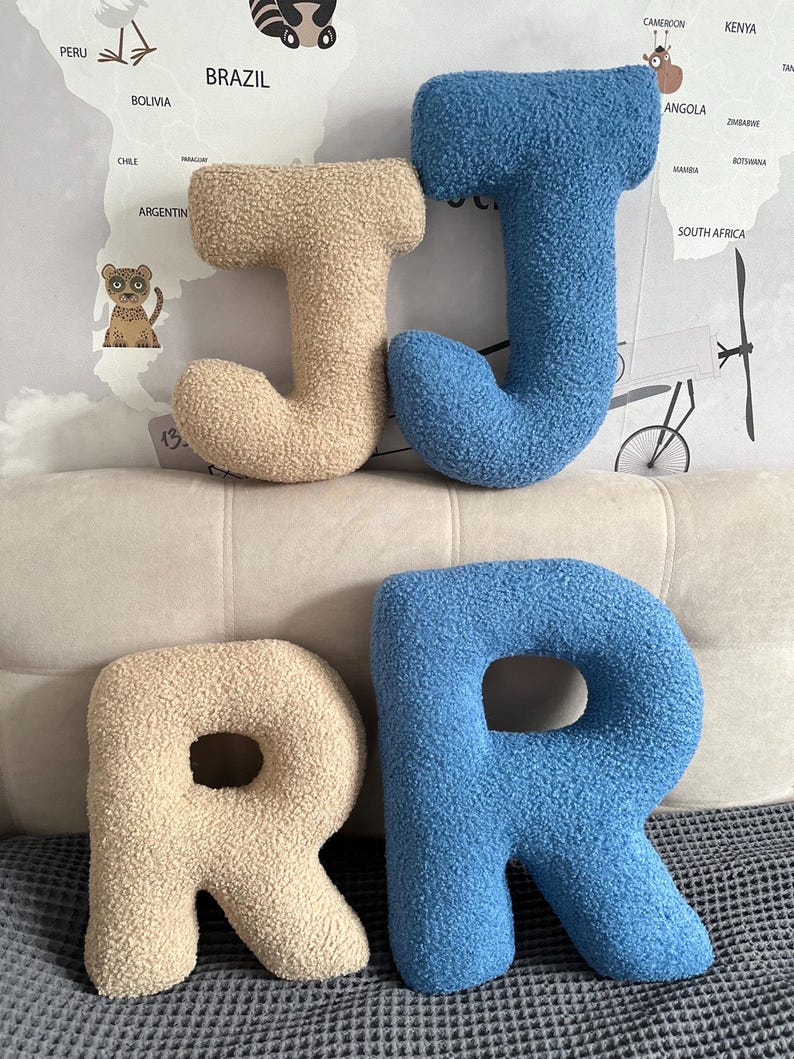 K&ouml;nnte beinhalten: Vier Buchstabenkissen aus strukturiertem, sherpa-artigem Stoff. Zwei Kissen haben die Form des Buchstabens J, eines in Beige und eines in Blau. Zwei Kissen haben die Form des Buchstabens R, eines in Beige und eines in Blau. Weiche, abgerundete Form.