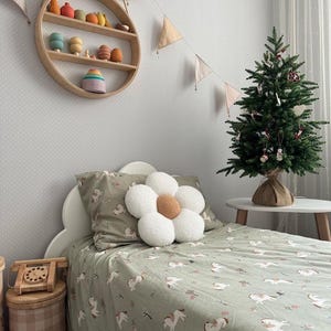 May include: A child's bedroom with a bed featuring a green unicorn-patterned duvet cover and pillowcase. A large white flower-shaped pillow sits on top. A small Christmas tree and wooden toys decorate the room. A round wooden shelf hangs on the wall.