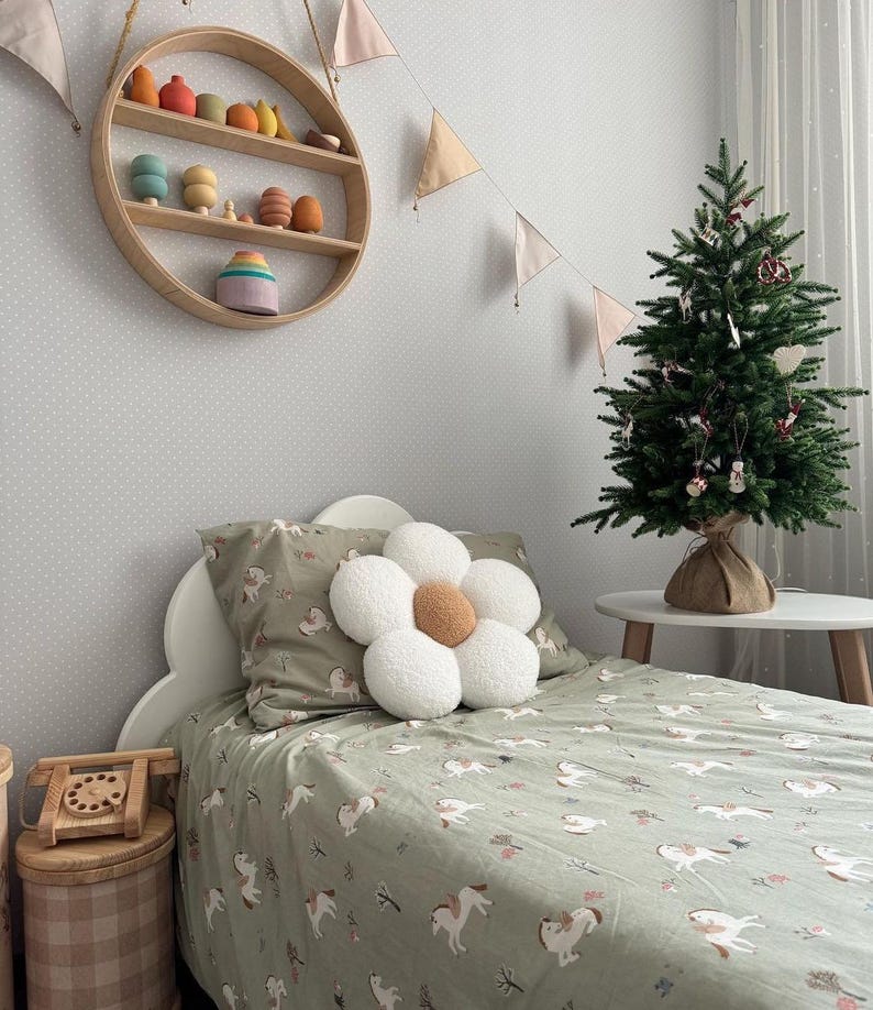 May include: A child's bedroom with a bed featuring a green unicorn-patterned duvet cover and pillowcase. A large white flower-shaped pillow sits on top. A small Christmas tree and wooden toys decorate the room. A round wooden shelf hangs on the wall.