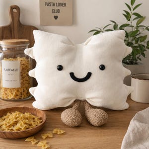 Pode incluir: Uma almofada de pelúcia cor de creme em forma de laço de massa farfalle, com dois pequenos olhos pretos bordados e uma boca preta curva. A almofada tem dois pés arredondados e texturizados em castanho claro na parte inferior.