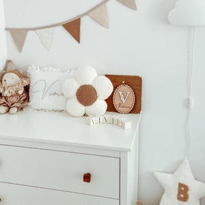 May include: A white dresser with wooden knobs holds a flower-shaped pillow with a brown center, a pillow with the name "Vivie" embroidered on it, and wooden blocks spelling "VIVIEN". A star-shaped pillow with the letter "B" is also present.