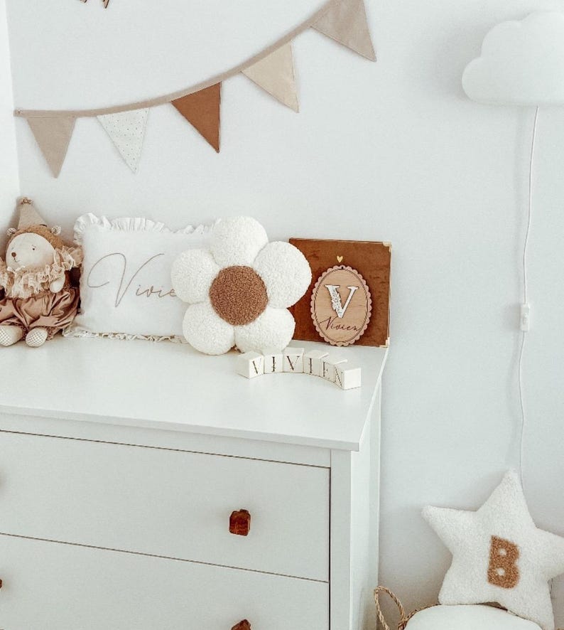 May include: A white dresser with wooden knobs holds a flower-shaped pillow with a brown center, a pillow with the name "Vivie" embroidered on it, and wooden blocks spelling "VIVIEN". A star-shaped pillow with the letter "B" is also present.