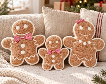 May include: Three gingerbread man-shaped plush cushions made of textured brown fabric with white icing details. Two cushions feature red and white striped bow ties, while the third has a small red and white striped hair bow and white icing necklace.