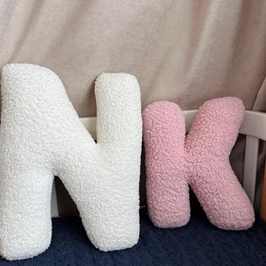 K&ouml;nnte beinhalten: Zwei pl&uuml;schige buchstabenf&ouml;rmige Kissen aus wei&szlig;em und rosa strukturiertem Sherpa-Stoff. Das gro&szlig;e wei&szlig;e Kissen hat die Form des Buchstabens N und das kleinere rosa Kissen die Form des Kleinbuchstabens k, beide mit abgerundeten Kanten.