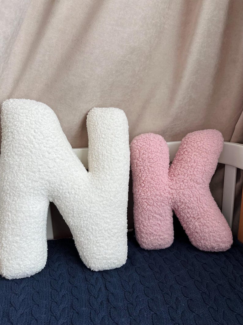 K&ouml;nnte beinhalten: Zwei pl&uuml;schige buchstabenf&ouml;rmige Kissen aus wei&szlig;em und rosa strukturiertem Sherpa-Stoff. Das gro&szlig;e wei&szlig;e Kissen hat die Form des Buchstabens N und das kleinere rosa Kissen die Form des Kleinbuchstabens k, beide mit abgerundeten Kanten.