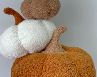 May include: A stack of three plush pumpkins in autumnal colors. The bottom pumpkin is orange, the middle one is white, and the top one is brown. Each pumpkin has a textured, fuzzy surface and a curved stem. A decorative fall-themed item.