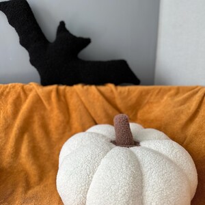 May include: A white plush pumpkin pillow with a brown stem. The pillow is sitting on a brown blanket.
