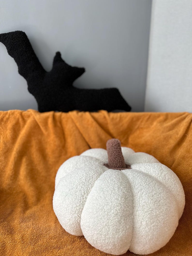 May include: A white plush pumpkin pillow with a brown stem. The pillow is sitting on a brown blanket.