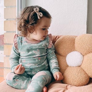 May include: A child's two-piece pajama set in a light teal color with a bunny print. The long-sleeved top has pink ruffles and trim. The matching pants have pink knee patches. A beige bow hair clip is in the child's hair.