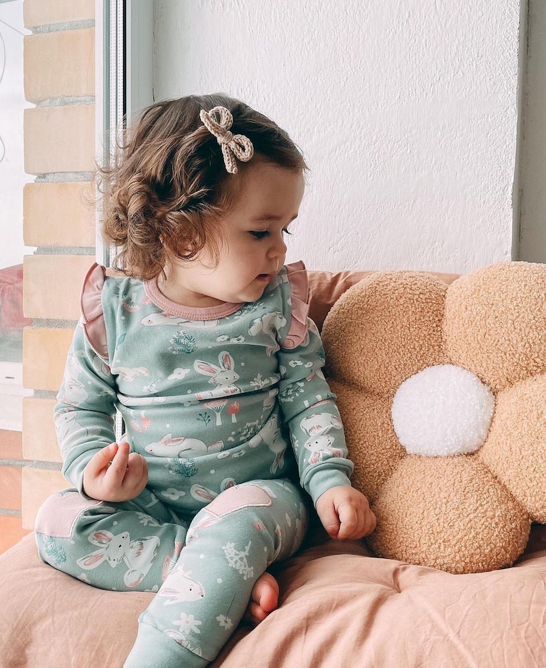 May include: A child's two-piece pajama set in a light teal color with a bunny print. The long-sleeved top has pink ruffles and trim. The matching pants have pink knee patches. A beige bow hair clip is in the child's hair.