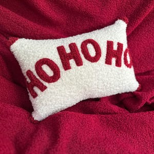 Può includere: Un cuscino rettangolare bianco con la scritta "HOHOHO" in rosso, posto su una superficie rossa strutturata. Il cuscino ha una consistenza soffice e bordi rossi. Un oggetto decorativo festivo.