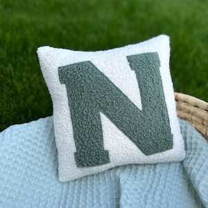 Könnte beinhalten: Ein quadratisches, weißes, strukturiertes Kissen mit einem großen, dunkelgrünen Buchstaben "N" in der Mitte. Das Kissen liegt auf einer hellblauen Decke und einem Weidenkorb, vor einem grünen, grasbewachsenen Hintergrund.