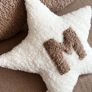 以下が含まれることがあります： 中央に茶色のふわふわした文字「M」がついた白い星型のクッション。クッションは柔らかくふわふわした素材でできています。