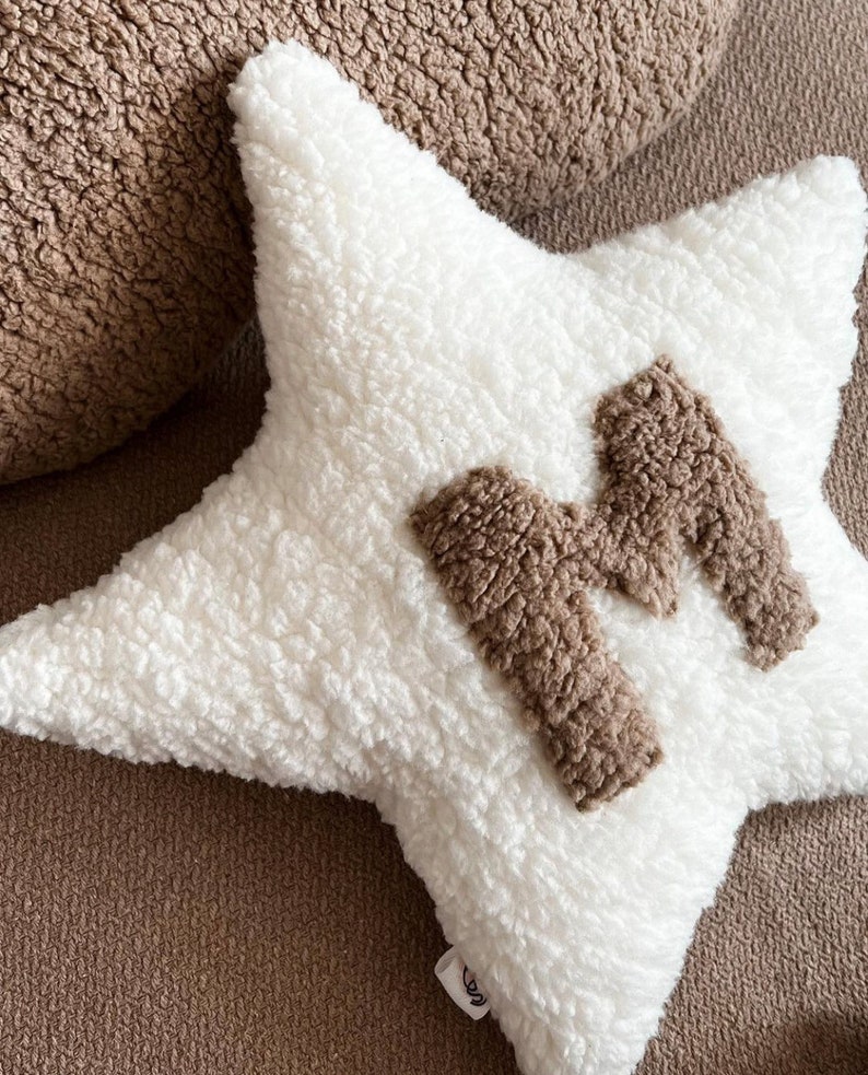 以下が含まれることがあります： 中央に茶色のふわふわした文字「M」がついた白い星型のクッション。クッションは柔らかくふわふわした素材でできています。