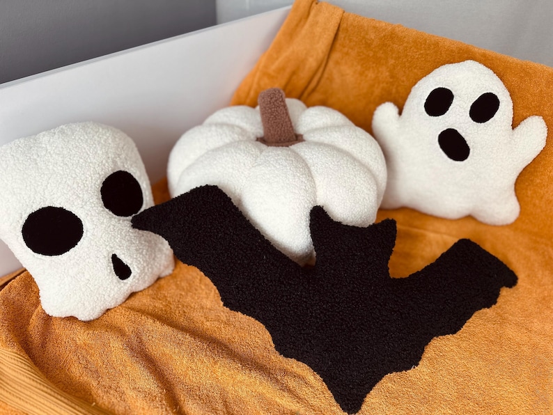 May include: A set of five Halloween-themed plush pillows. The pillows are shaped like a ghost, a pumpkin, a skull, and two bats. The pillows are all white with black accents.