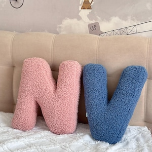 K&ouml;nnte beinhalten: Zwei buchstabenf&ouml;rmige Kissen aus strukturiertem Sherpa-Stoff. Ein Kissen ist in Form des Buchstabens N in einem zarten Rosaton gehalten, das andere in Form des Buchstabens V in einem gedeckten Blauton. Beide Kissen haben eine dicke, lockige Fleece-Struktur.