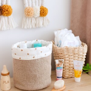 Peut inclure: Un panier de rangement en tissu marron avec un dessus en tissu blanc orné de petits cœurs dorés. Le panier est rempli d'articles de soin pour bébé, dont une bouteille de lotion, un tube de crème et une petite bouteille en bois étiquetée "Lait". Il y a aussi un peigne et une brosse en bois, ainsi qu'un panier en osier rempli de couches.