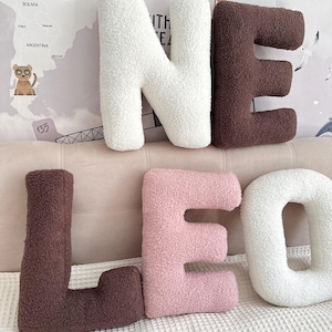K&ouml;nnte beinhalten: Eine Ansammlung von sechs buchstabenf&ouml;rmigen Kissen aus strukturiertem Sherpa-Stoff. Die Buchstaben bilden die W&ouml;rter N, E, L, E, O und O in einer Farbpalette aus Creme, Dunkelbraun und zartem Rosa, angeordnet in zwei Reihen auf einer hellen Fl&auml;che.
