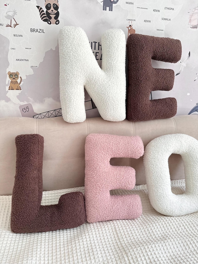 K&ouml;nnte beinhalten: Eine Ansammlung von sechs buchstabenf&ouml;rmigen Kissen aus strukturiertem Sherpa-Stoff. Die Buchstaben bilden die W&ouml;rter N, E, L, E, O und O in einer Farbpalette aus Creme, Dunkelbraun und zartem Rosa, angeordnet in zwei Reihen auf einer hellen Fl&auml;che.