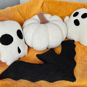 May include: A set of four Halloween-themed plush pillows. The pillows are shaped like a pumpkin, a ghost, a bat, and a skull. The pillows are all white with black accents. The pillows are sitting on a yellow blanket.