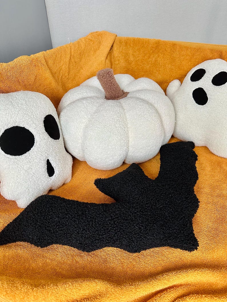 May include: A set of four Halloween-themed plush pillows. The pillows are shaped like a pumpkin, a ghost, a bat, and a skull. The pillows are all white with black accents. The pillows are sitting on a yellow blanket.