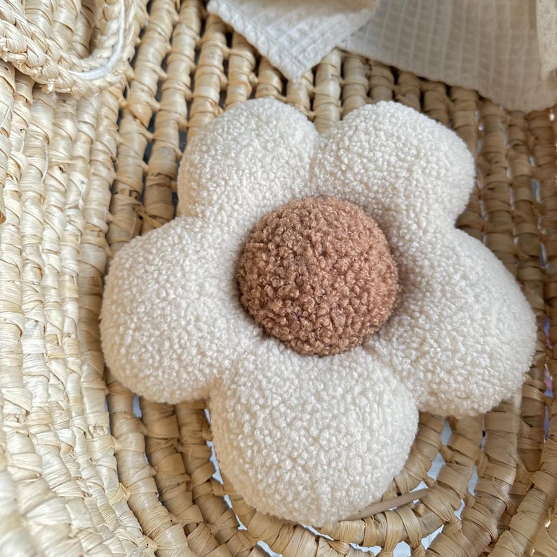 May include: A plush, flower-shaped pillow with a cream-colored, textured exterior and a brown center. The pillow is made of a soft, fuzzy material and is placed on a woven basket. The flower pillow adds a touch of whimsy to any room.