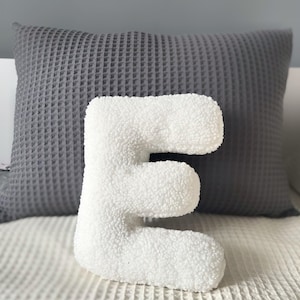K&ouml;nnte beinhalten: Ein wei&szlig;es, pl&uuml;schiges, strukturiertes Kissen in Form des Buchstabens E mit einer weichen, flauschigen Oberfl&auml;che. Der Buchstabe ist in einer abgerundeten Blockschrift gestaltet und steht aufrecht, was eine dicke, gepolsterte Optik bietet.