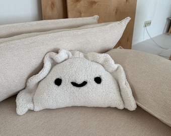 May include: A white plush pillow shaped like a dumpling with a black smiling face. The pillow is sitting on a beige couch.