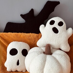 May include: Three plush Halloween-themed pillows: a white ghost with black eyes and a mouth, a white pumpkin with a brown stem, and a black bat with outstretched wings.
