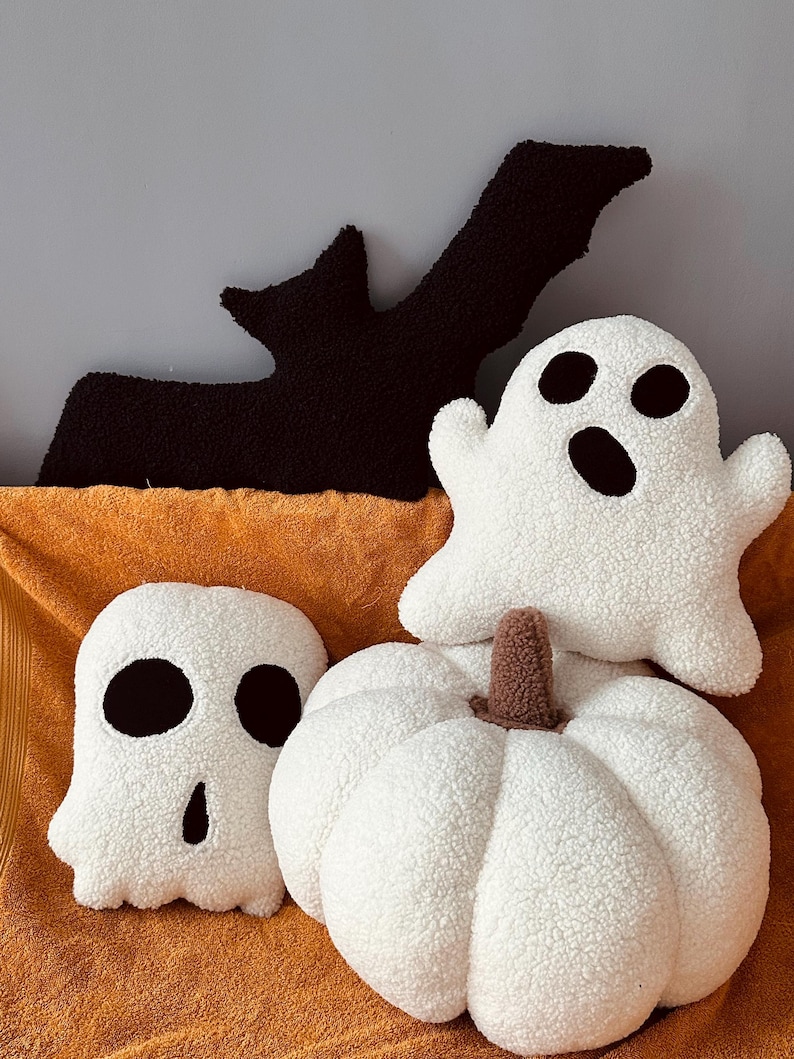 May include: Three plush Halloween-themed pillows: a white ghost with black eyes and a mouth, a white pumpkin with a brown stem, and a black bat with outstretched wings.